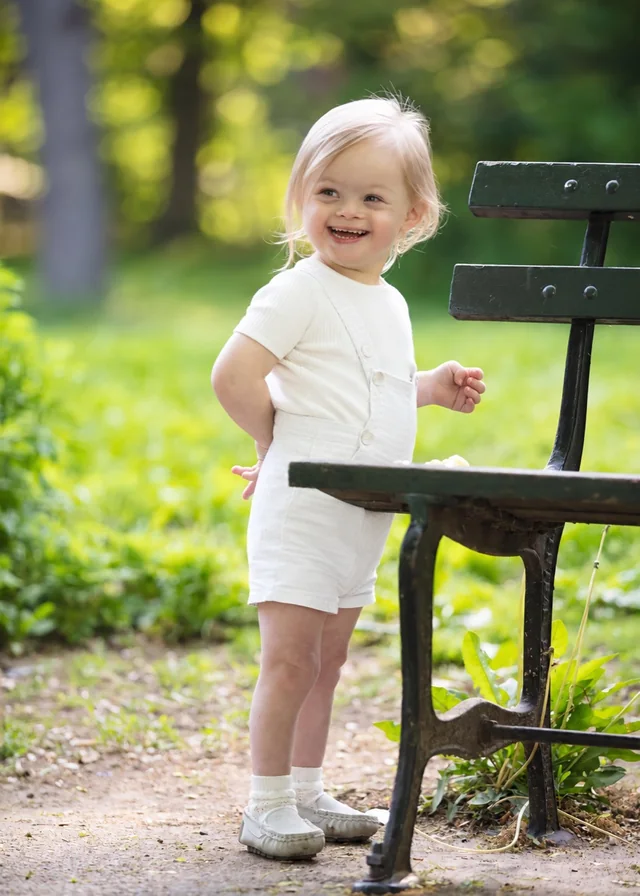 A smiling toddler with Down syndrome standing by a park bench outdoors