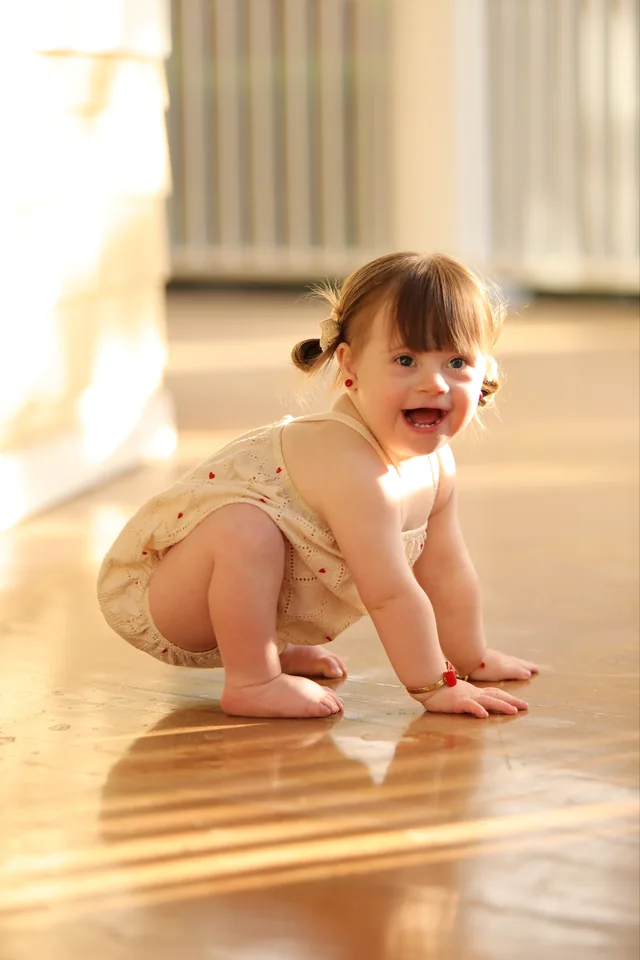 A smiling toddler in a cream outfit, playing on a sunlit wooden floor